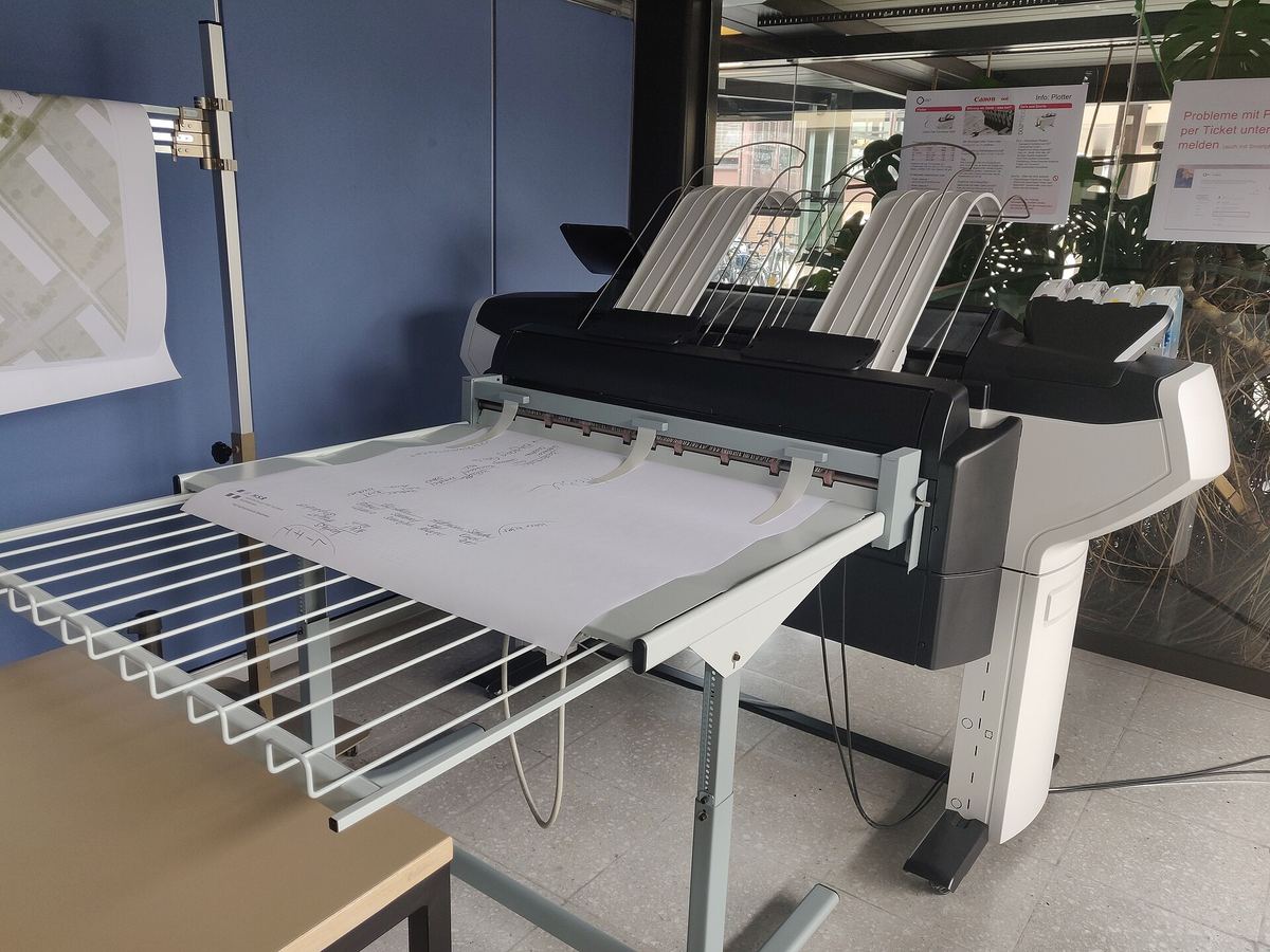 Großformatiger Plotter in einem Hochschulraum mit Ablagekorb, auf dem ein ausgedruckter Plan liegt